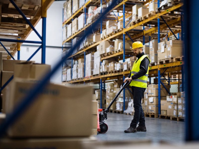 Young male warehouse worker pulling a pallet truck with boxes.